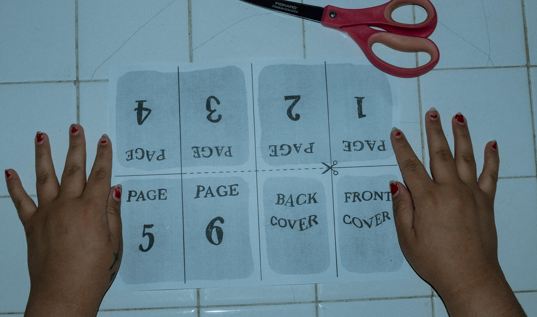 A grainy, blue-toned photo of an 8 page mini zine before being folding with two hands resting on each side of the frame and a pair on scissors on a tile table.